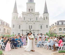 Bride and groom kiss outdoors before a white historic building as guests clap and smile at their wedding ceremony.