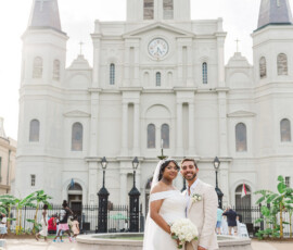 Bride and groom smile in wedding attire before St. Louis Cathedral, New Orleans, with towers and clock in the background.