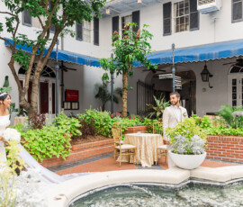 Bride and groom smile in a lush courtyard with greenery, fountain, and table settings under a blue awning at a Gulf South wedding.