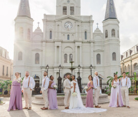 Wedding party in front of white church with tall spires; bride, groom, bridesmaids in lavender, groomsmen in beige.