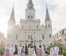 Wedding party in pastels poses smiling before a grand three-spired New Orleans cathedral. Bride and groom stand centered.