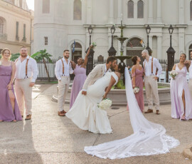 Bride and groom kiss as he dips her, wedding party in lavender and suspenders pose joyfully outside a historic New Orleans building.