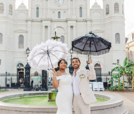 Newlyweds with decorative umbrellas pose by a fountain outside a historic New Orleans church, smiling in wedding attire.