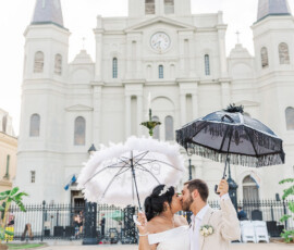 Bride and groom kiss with decorative umbrellas by St. Louis Cathedral, New Orleans, near a green fountain.