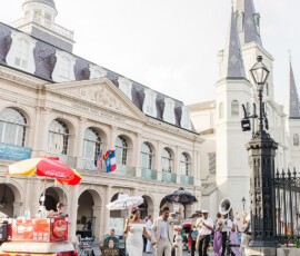 Bride and groom with parasols stroll by street band and hot dog stand near Louisiana State Museum, St. Louis Cathedral in view.