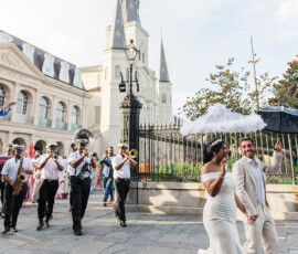 Bride and groom lead a joyful New Orleans Second Line wedding parade with brass band, white umbrella, and smiles outside a church.