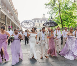 Wedding party in pastel attire walks joyfully through New Orleans with bouquets, parasols, historic buildings, and trees behind them.