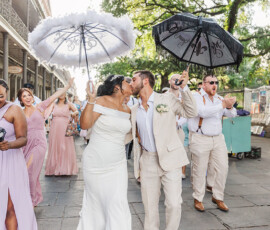 A newlywed couple kisses and leads a festive Gulf South wedding parade with decorated umbrellas as pastel-clad guests celebrate.
