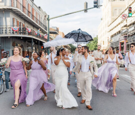 A joyful wedding party in pastels crosses a sunny New Orleans street, bride with white parasol, groom with black.