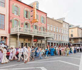 Colorful parade inspired by New Orleans, people march past historic buildings and a SINGAPORE sign as spectators enjoy the festivities.