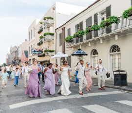 Bride and groom with umbrellas lead a pastel-clad wedding party down a city street, cheered on by smiling bystanders.
