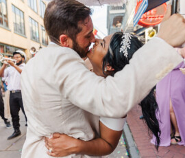 Smiling couple embraces on a lively New Orleans street wedding, with brass musicians; bride in white, groom in beige suit.