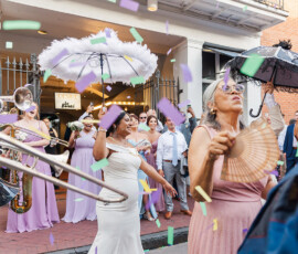 Louisiana wedding parade with musicians, pastel-clad guests, confetti, fans, and decorative umbrellas on a city street.