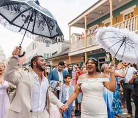 A joyful wedding couple dances in the street with decorated umbrellas, cheered by guests and colorful historic Louisiana buildings.