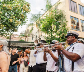 Brass band in white uniforms plays trumpets outdoors, entertaining a joyful crowd with lively, festive street music.