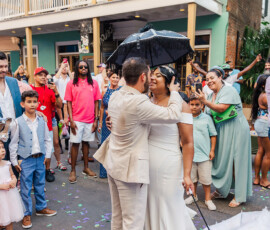 Newlyweds kiss under a black umbrella at a lively Gulf South wedding, with confetti, smiling guests, and a mint-green building.