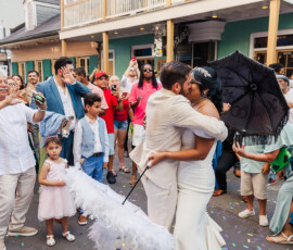 Bride and groom kiss on a confetti-strewn New Orleans street, surrounded by cheering guests and colorful buildings.