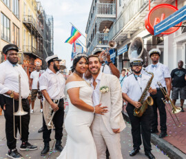 Bride and groom embrace on a city street with a brass band and rainbow flag, celebrating a vibrant New Orleans wedding.