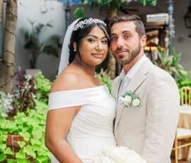 Bride in off-shoulder white dress with roses stands by groom in beige suit, outdoors with greenery and lights at Louisiana wedding.