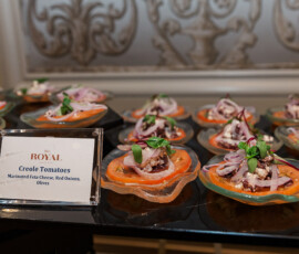Small plates of sliced tomatoes with feta, onions, olives, and microgreens sit by a Creole Tomatoes sign; ornate wallpaper behind.