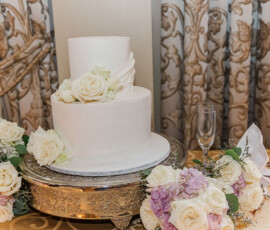 Elegant white two-tiered wedding cake with roses on a silver stand, surrounded by flowers and classic patterned curtains.