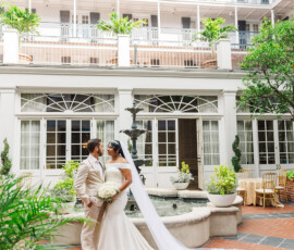 Bride in long veil with bouquet and groom in light suit by courtyard fountain, elegant New Orleans wedding setting.