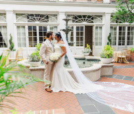 Bride in off-shoulder gown and groom in light suit stand by a fountain at their joyful New Orleans wedding.