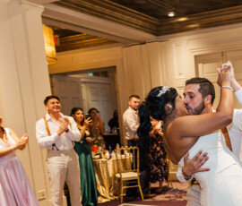 Newlyweds kiss during their first dance at a Louisiana wedding, surrounded by smiling, clapping guests in formal attire.