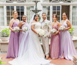 Bride, groom, and four bridesmaids in lilac dresses smile together outside a windowed building at their Gulf South wedding.