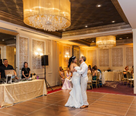 Bride and groom share their first dance in a chandelier-lit ballroom as guests watch and a DJ stands by his equipment.