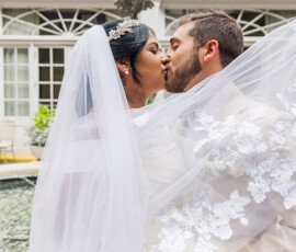Bride and groom kiss by a New Orleans fountain, wrapped in her veil. She holds white roses; both wear elegant wedding attire.