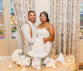 Bride and groom smile behind a flower-adorned cake table at their elegant Louisiana wedding, curtains and windows behind them.