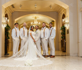 Bride and groom with five groomsmen in white pose under chandeliers; bouquet rests by bride’s train. Louisiana wedding inspiration.