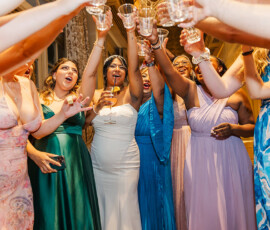 Women in colorful dresses, including a bride in white, toast at a festive indoor Louisiana wedding reception.