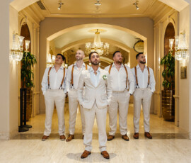 Five men in cream suits pose in an elegant hallway with chandeliers, capturing Louisiana wedding inspiration.