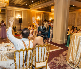 Bride and groom toast at a decorated table, surrounded by guests in an elegant, warmly lit wedding banquet hall.
