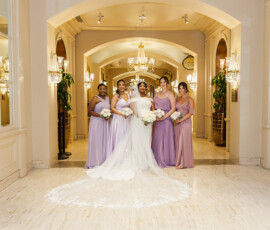 Bride in white gown with four bridesmaids in lavender, holding white bouquets in a chandelier-lit New Orleans wedding hallway.