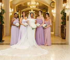 Bride in white and five women in pastel purple dresses pose indoors with bouquets, under chandeliers and arches at a formal wedding.