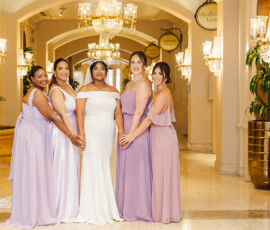 Bride in white and bridesmaids in lavender smile, holding hands under chandeliers at a Louisiana wedding hallway.
