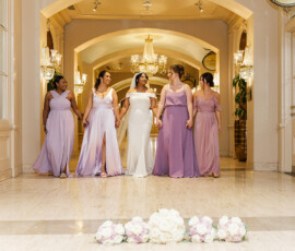 Bride in white and bridesmaids in lavender walk hand-in-hand under chandeliers, with a floral bouquet on the floor.
