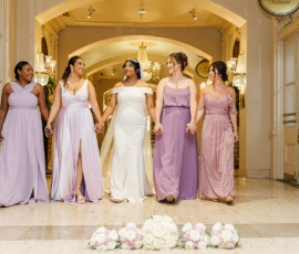 Bride and bridesmaids in lavender dresses walk hand in hand indoors, smiling by a bouquet after their Louisiana wedding.