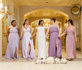 Five smiling women in formal dresses, including a bride, walk hand-in-hand indoors at a classic New Orleans wedding.