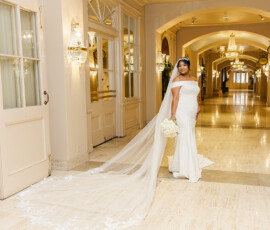 Bride in off-shoulder white gown and veil holds white rose bouquet indoors, with chandeliers and elegant New Orleans décor.