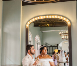 Couple in wedding attire holding champagne glasses under a chandelier in an elegant room with mirrors and arched windows.