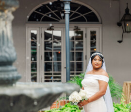 Bride in off-shoulder white dress with veil holds bouquet by a fountain, brick planter, and large windows—timeless wedding inspiration.