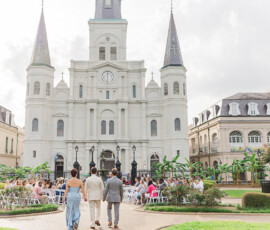 A couple walks down an outdoor aisle toward guests in front of a historic white cathedral with three towers in Louisiana.