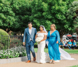 Bride in white gown with bouquet walks outdoors, joined by man and woman in blue, trees and guests behind—Gulf South wedding.
