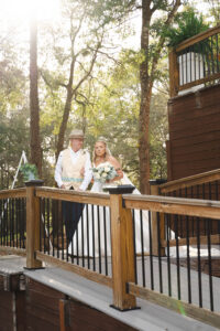 Bride in white dress and veil walks with older man down a wooden ramp outdoors, amid trees and sunlight, Gulf South wedding.