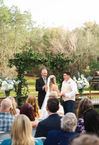 Bride and groom hold hands with officiant under leafy arch at Louisiana wedding, surrounded by guests, trees, and floral decor.