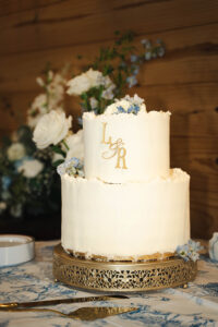 Elegant two-tier white wedding cake with gold L&R topper, blue flowers, and gold stand, set against floral and wood backdrop.